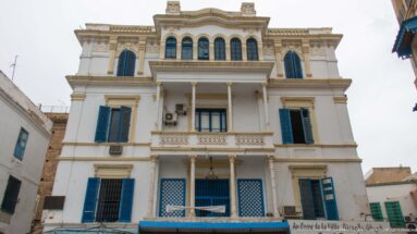 White Tunis building with blue shutters, columned balconies, arched upper windows, and decorative parapet.