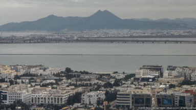 Elevated view across Tunis neighborhoods and the Lake of Tunis toward a dark mountain ridge under gray skies.