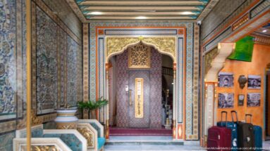 Ornate hotel passage with patterned tile walls, painted borders, carved arches, and a decorated doorway.
