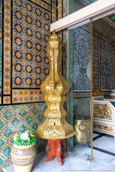 Tall pierced brass decorative object beside a ceramic planter against richly patterned blue and gold tile walls.