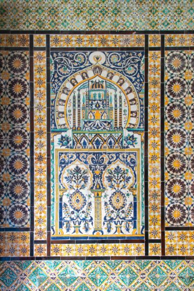 Blue, green, and ochre tile panel with stylized architecture, floral motifs, and patterned borders.