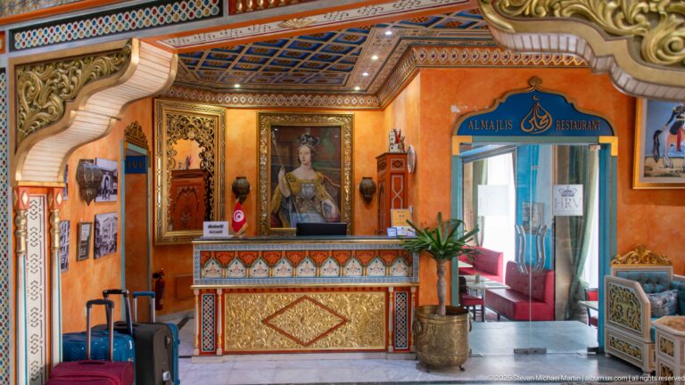 Ornate hotel reception with patterned desk, framed portrait, luggage, and an entrance marked Al Majlis Restaurant.