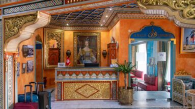 Ornate hotel reception with patterned desk, framed portrait, luggage, and an entrance marked Al Majlis Restaurant.