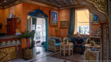Guest seated in an ornate hotel lounge beside reception, patterned furniture, carved arches, and Al Majlis Restaurant.