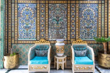 Two turquoise armchairs flank a painted vase against richly patterned ceramic wall panels in a Tunis hotel.