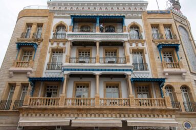 Upward view of the Hotel Royal Victoria façade with stacked balconies, arched openings, and blue-painted awnings.