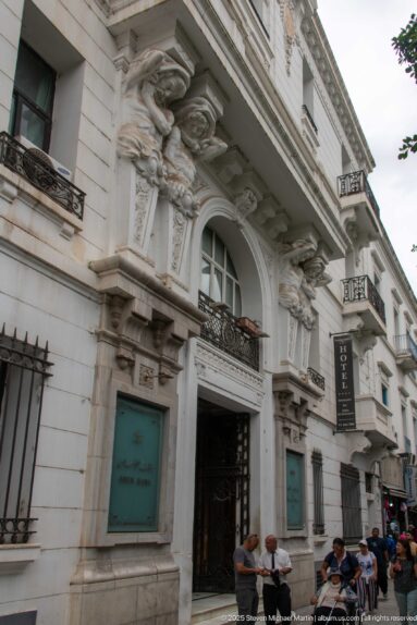 Ornate white Tunis façade with balconies, arched windows, sculptural brackets, and pedestrians at street level.