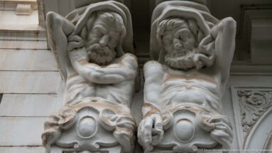 Paired sculpted bearded male figures with bent arms projecting from a white building façade as decorative supports.