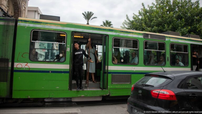 Green Tunis city bus stopped in traffic with open doors, visible passengers, and a dark car passing alongside.