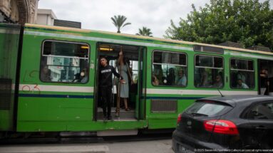 Green Tunis city bus stopped in traffic with open doors, visible passengers, and a dark car passing alongside.