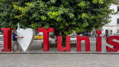 Large red I Love Tunis sign with a white heart beneath a tree and a person standing beside it.