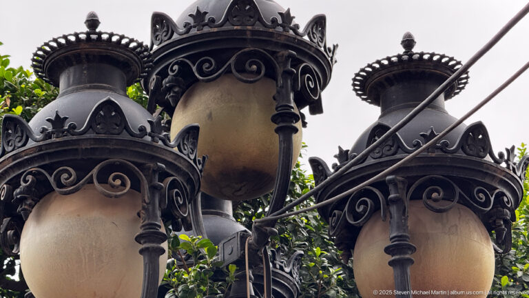 Low-angle close view of ornate black metal street lanterns with scrollwork, finials, and warm glass globes.