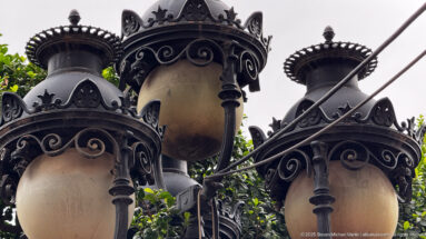 Low-angle close view of ornate black metal street lanterns with scrollwork, finials, and warm glass globes.