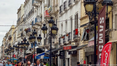 Busy Tunis street lined with pale façades, shops, pedestrians, and repeated ornate black multi-globe lamps.