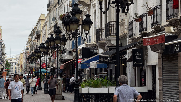 Crowded central Tunis street with cafés, balconies, hotel signs, and rows of ornate black lanterns.