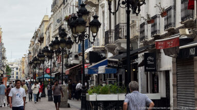 Crowded central Tunis street with cafés, balconies, hotel signs, and rows of ornate black lanterns.