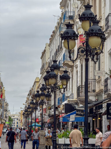Low-angle perspective of ornate multi-globe street lamps receding above pedestrians and cafés in central Tunis.