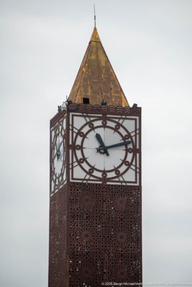 Close view of the Tunis Clock Tower clock face, pyramidal roof, and perforated reddish metal shaft.