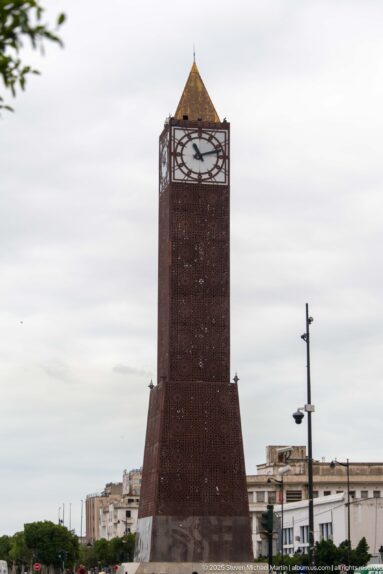 Full-height view of the reddish Tunis Clock Tower with flared base, perforated shaft, clock face, and pyramidal roof.