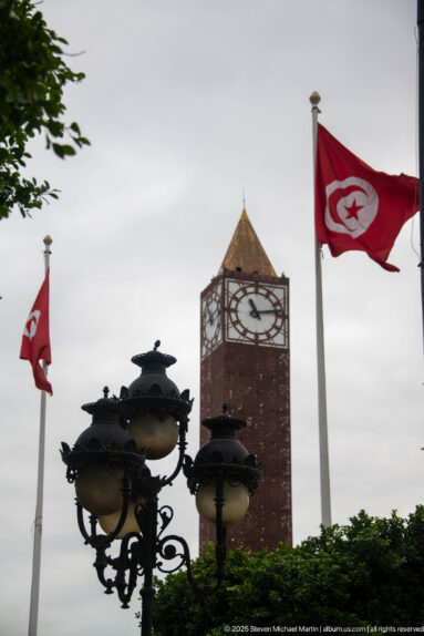 Tunis Clock Tower framed by red Tunisian flags, dark decorative lamps, and green hedges.