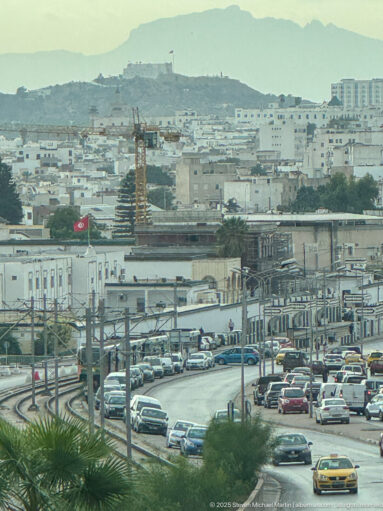 Elevated view over Tunis traffic, rail infrastructure, low-rise buildings, palm foliage, and a hazy ridge.