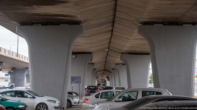 Cars parked beneath a concrete elevated roadway supported by repeated flared piers in Tunis.