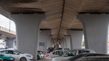 Cars parked beneath a concrete elevated roadway supported by repeated flared piers in Tunis.