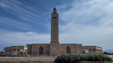 Frontal view of a large mosque with a tall square minaret, broad prayer hall, and arcaded side wings.