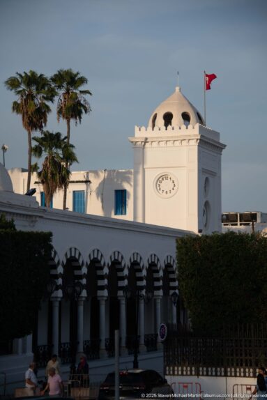 White domed clock pavilion with Tunisian flag above a long arcade of alternating dark and light arches.