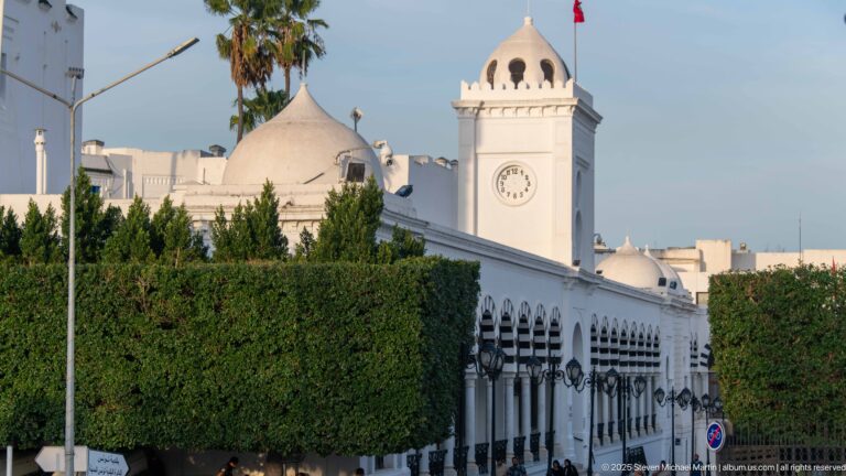 White domed clock pavilion with Tunisian flag above a long arcaded civic façade and clipped trees.
