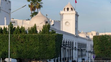 White domed clock pavilion with Tunisian flag above a long arcaded civic façade and clipped trees.