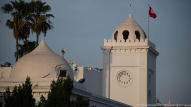 Close view of a white clock pavilion with circular dial, open dome, crenellated parapet, and Tunisian flag.