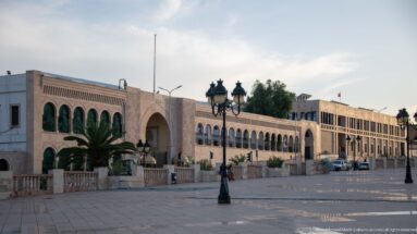 Long beige civic façade with repeated arches, green openings, palms, and ornate lamps beside a broad plaza.