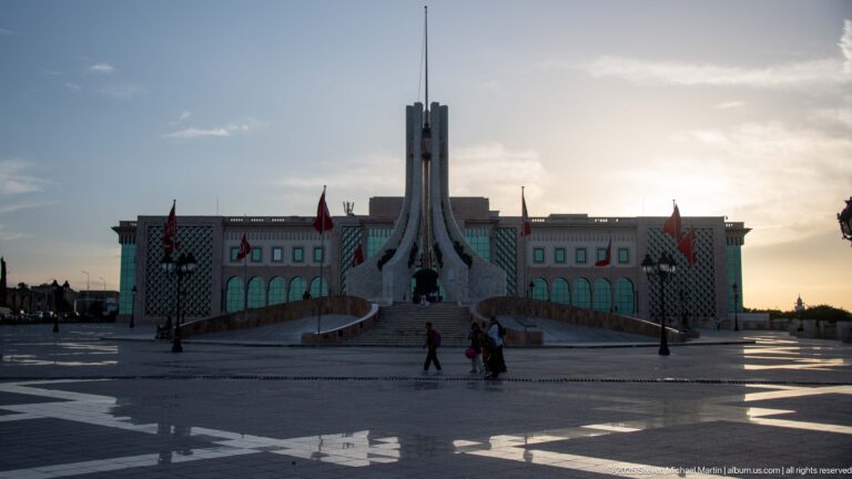 Tall pale monument on a stepped base in Kasbah Square with flags, pedestrians, and an administrative building behind.