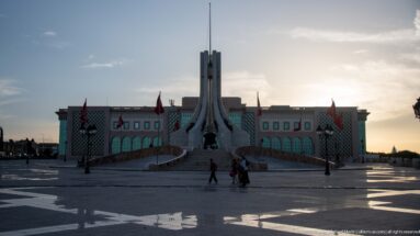 Tall pale monument on a stepped base in Kasbah Square with flags, pedestrians, and an administrative building behind.