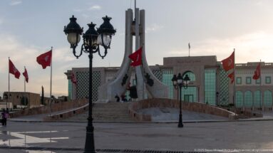 Closer view of the Kasbah Square monument with broad steps, curved ramps, Tunisian flags, lamps, and government building.