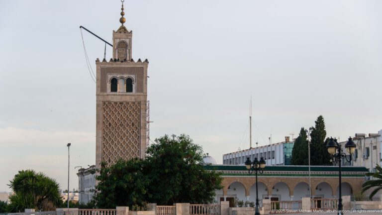 Kasbah Mosque minaret with diamond-pattern lattice rising above trees, arcades, and low white buildings.