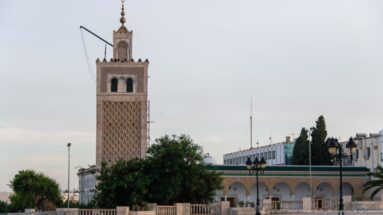 Kasbah Mosque minaret with diamond-pattern lattice rising above trees, arcades, and low white buildings.