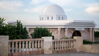 White hemispherical dome above a low pale building with shallow arches, recessed entrance, trees, and stone balustrade.