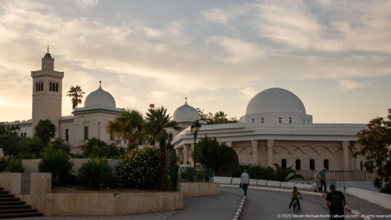 Kasbah Mosque minaret and several white domes rising above palms and pedestrians on a curving road at sunset.