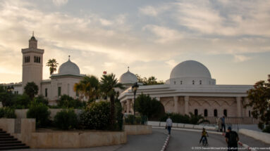 Kasbah Mosque minaret and several white domes rising above palms and pedestrians on a curving road at sunset.