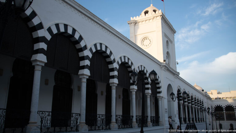 Oblique view along striped arches and iron railings toward the Ministry of Finance clock tower in Tunis.