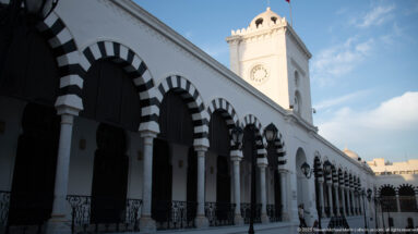 Oblique view along striped arches and iron railings toward the Ministry of Finance clock tower in Tunis.