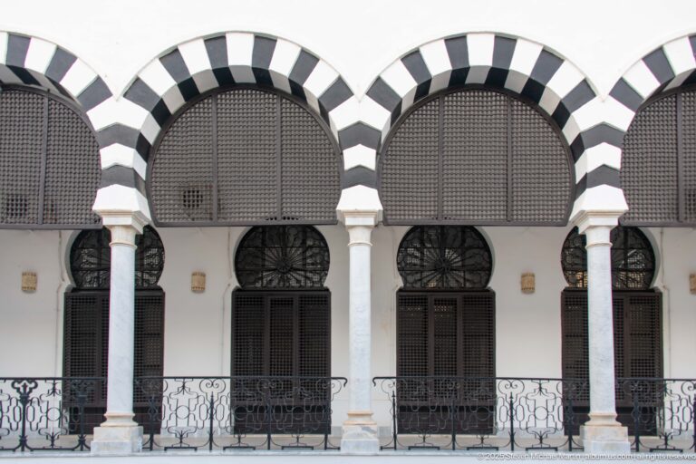 Three striped arches with white columns, dark screened openings, and wrought-iron railing on the Ministry of Finance façade.