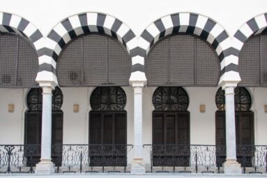 Three striped arches with white columns, dark screened openings, and wrought-iron railing on the Ministry of Finance façade.