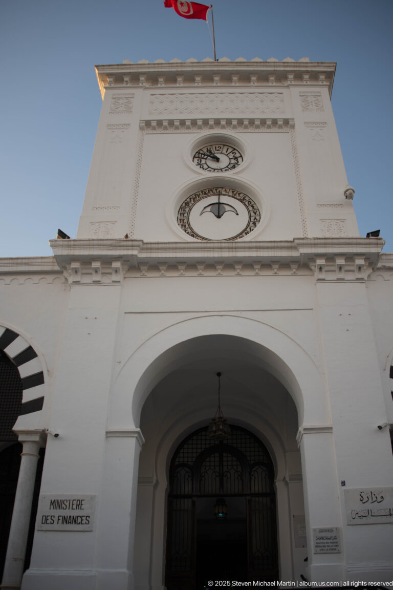 Main arched entrance of Tunisia's Ministry of Finance with clocks, Tunisian flag, and bilingual signage.