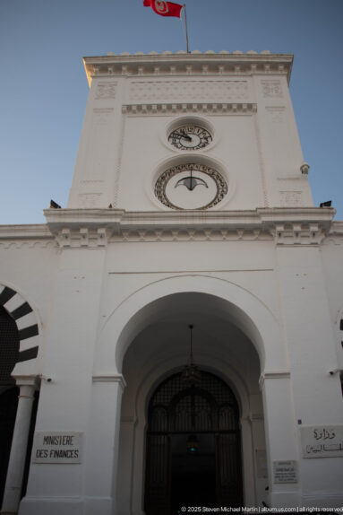 Main arched entrance of Tunisia's Ministry of Finance with clocks, Tunisian flag, and bilingual signage.