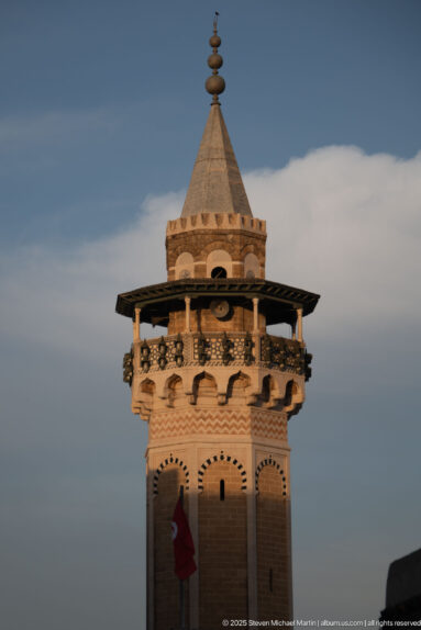 Ornate Tunis mosque minaret with projecting wooden gallery, patterned masonry, pyramidal roof, and stacked finial.