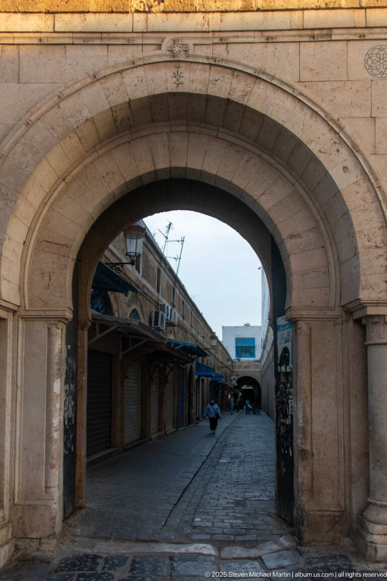Large stone arch opening onto a narrow Medina of Tunis lane with closed shops and covered stalls.