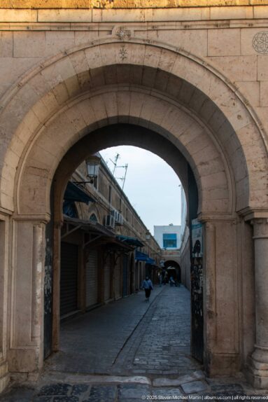 Large stone arch opening onto a narrow Medina of Tunis lane with closed shops and covered stalls.